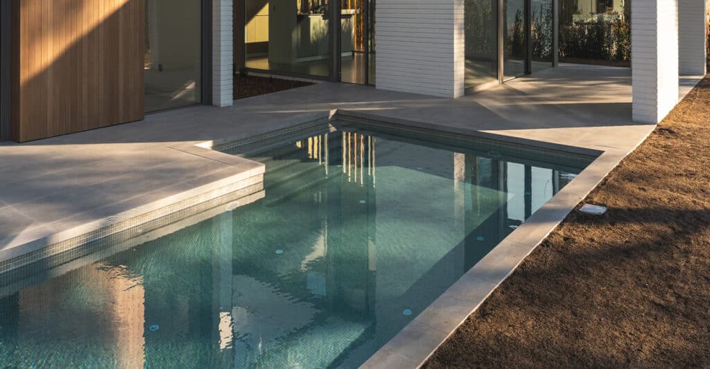 Piscine moderne à mosaïque verte près d'une maison contemporaine avec terrasse en béton clair et mur en bois. Lumière du soleil créant des reflets dans l'eau.