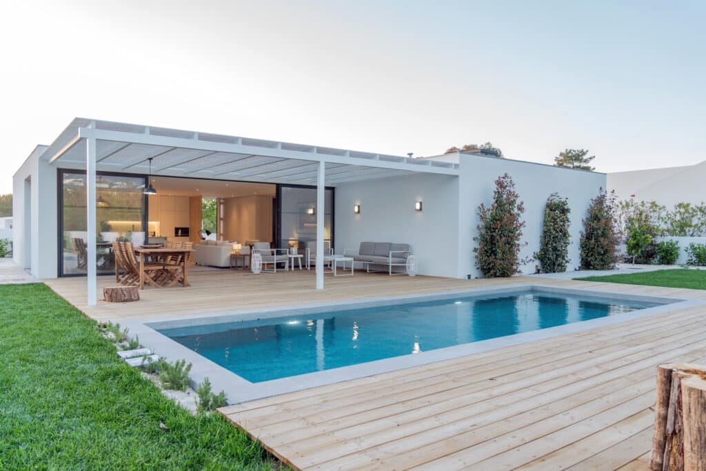 Maison moderne avec piscine, grande terrasse en bois, et pergola. Coin repas et salon extérieur. Jardin verdoyant attenant.