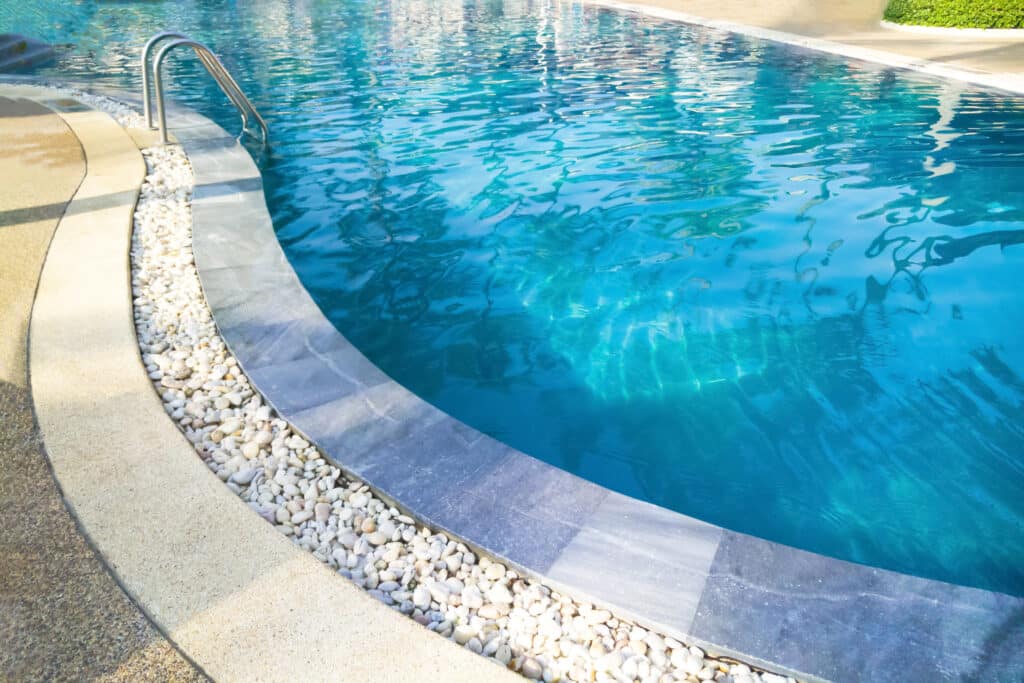Piscine bleue incurvée avec main courante, bordée de galets blancs et d'une terrasse beige. L'eau claire reflète la lumière du soleil.