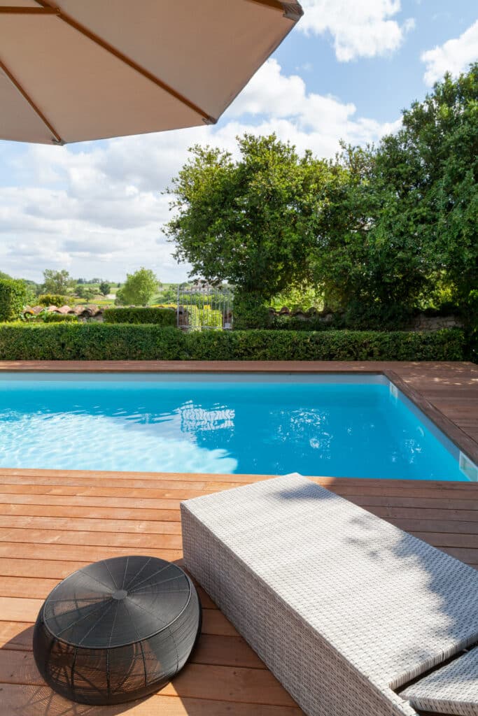 Piscine moderne avec terrasse en bois, transat et table d'extérieur sous parasol. Jardin luxuriant et campagne sous ciel bleu.